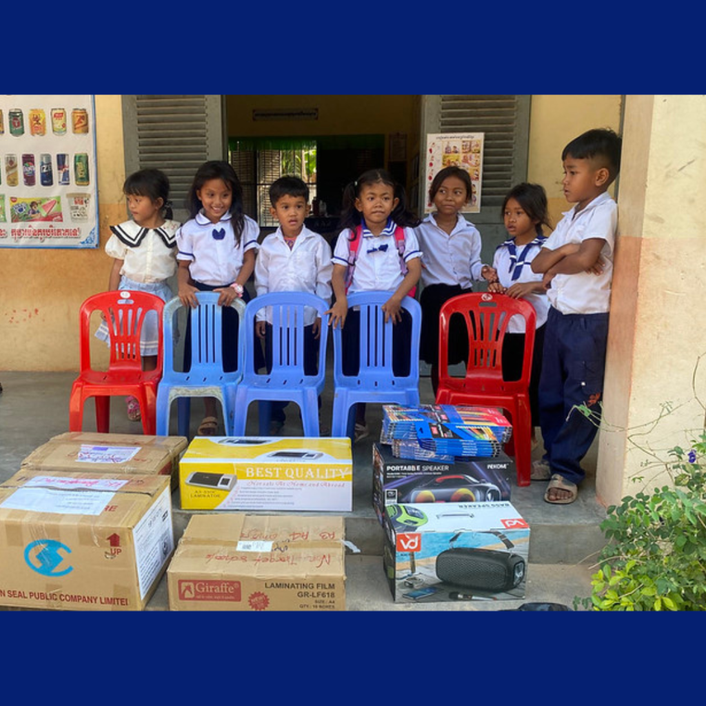Preschool children in uniform sit on plastic chairs behind boxes of newly delivered speakers and laminators