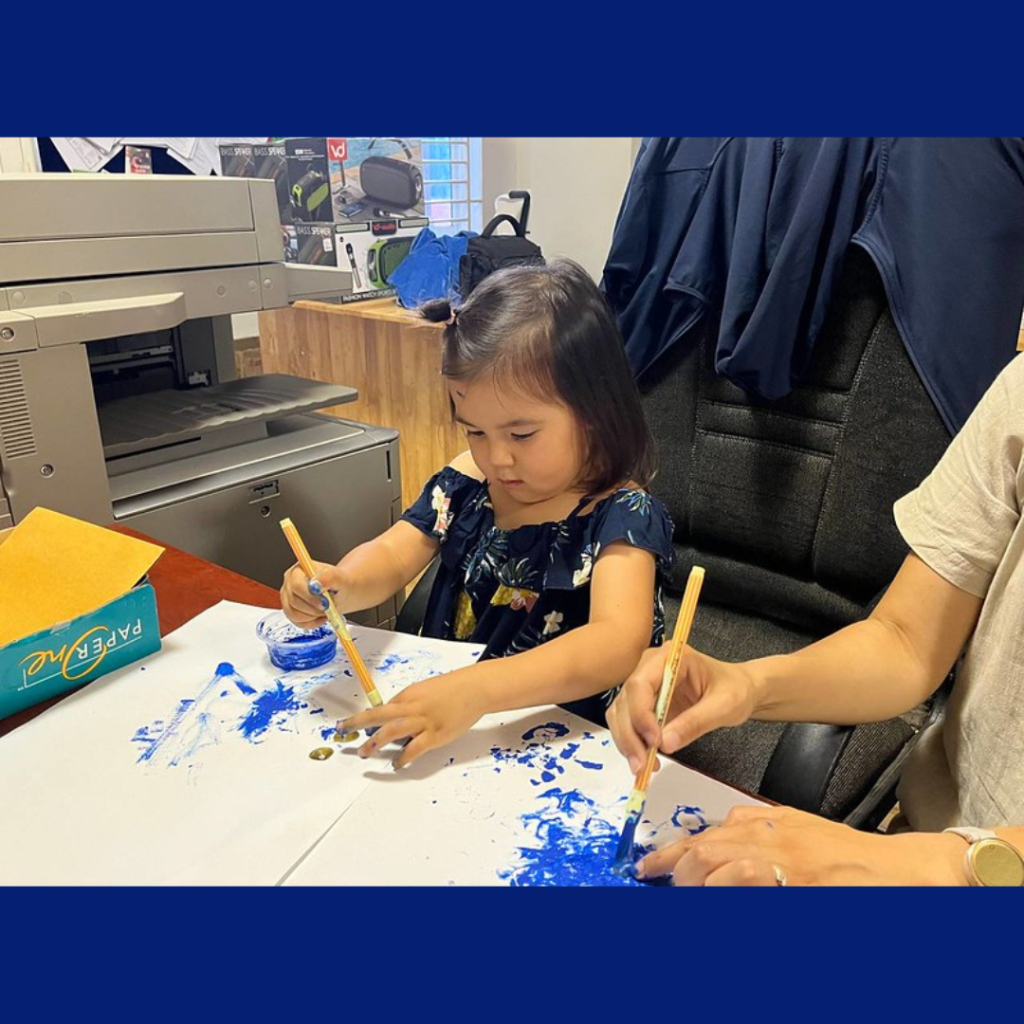 A toddler paints with a brush and blue paint at a table in the office, beside an adult's hand