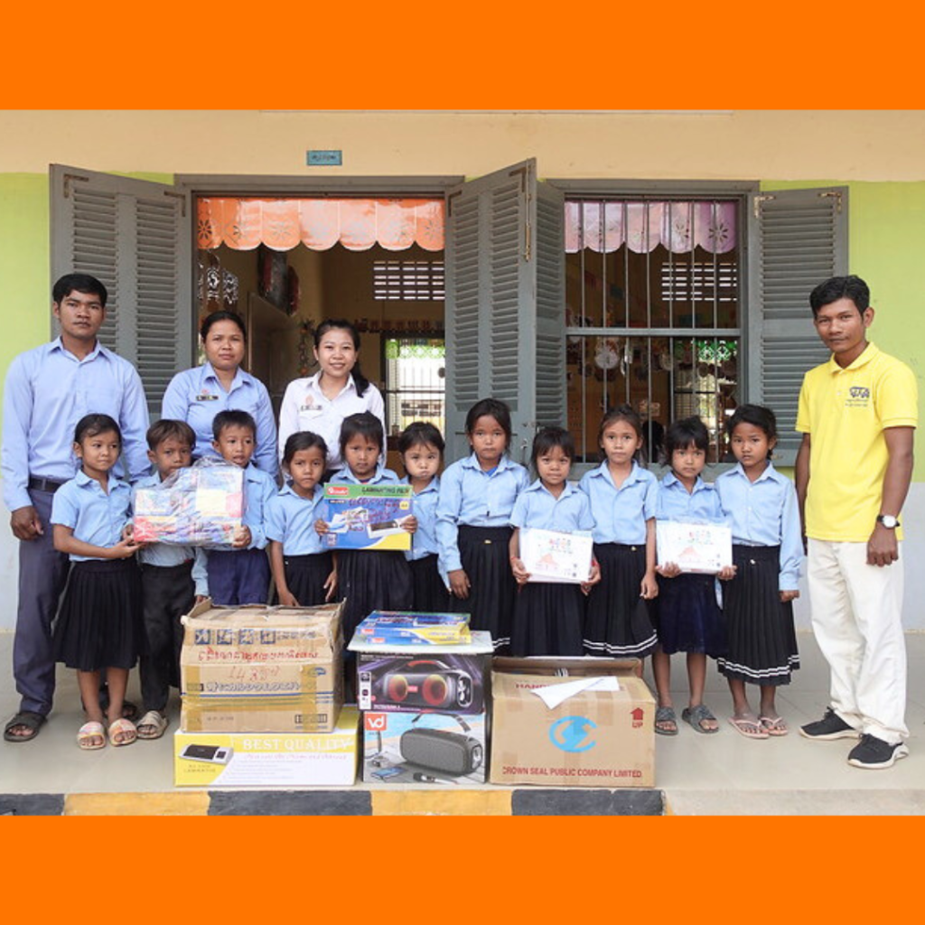 Preschool children in uniform, teachers and a TukTuk staff member stand behind boxes of delivered CLIP3 equipment