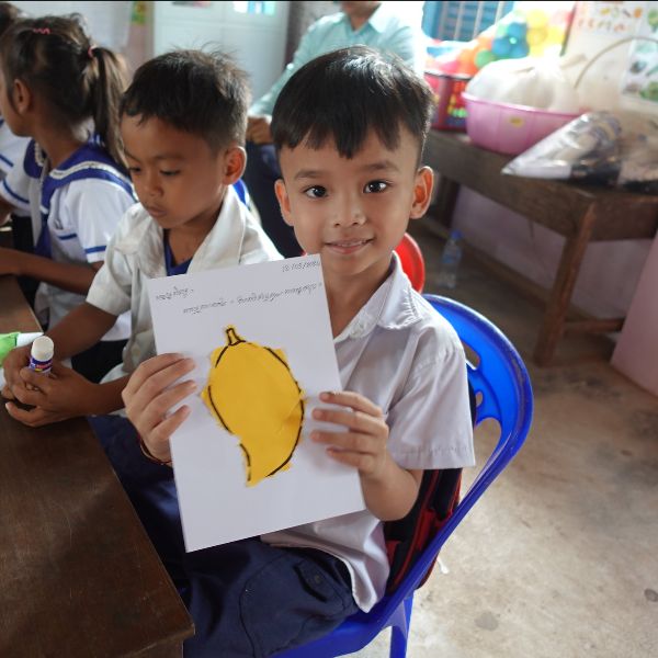 A smiling preschool boy in school uniform holds up a paper cut-out of a yellow fruit at his desk