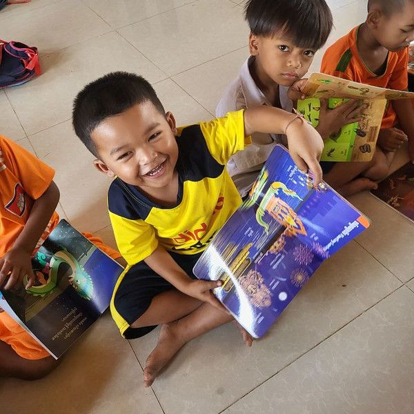 A smiling boy in a yellow shirt shows an open picture book while other children read on the floor