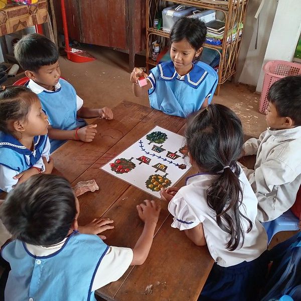 Preschool children in blue aprons and uniforms sit around a wooden table playing a tree picture matching game