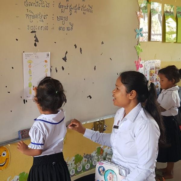 A preschool teacher in a white blouse and a small girl at a classroom wall chart match fruit pictures