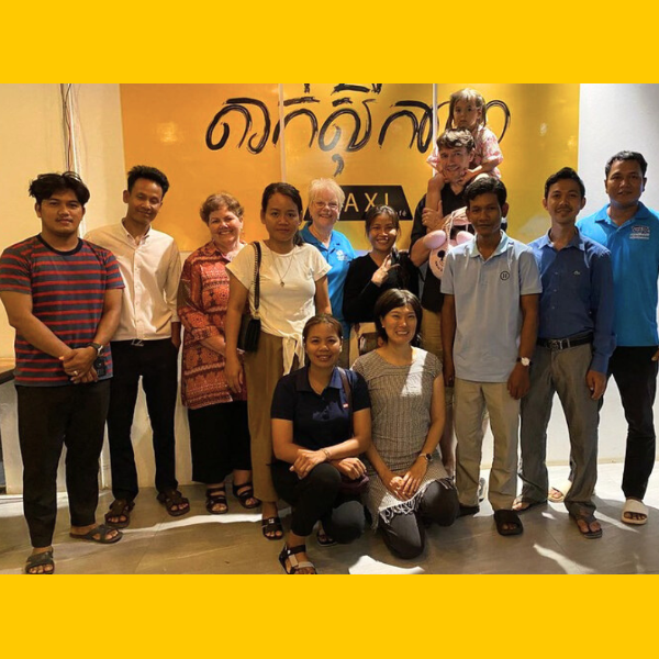A group of adults and a toddler pose together indoors in front of a yellow wall with Khmer lettering