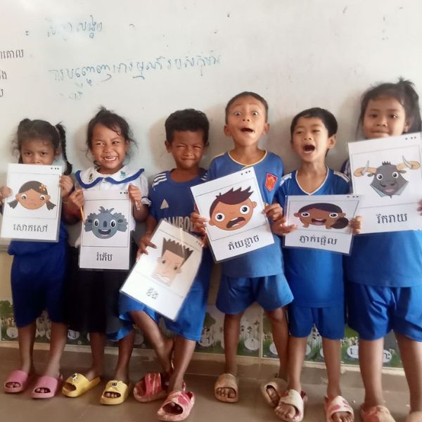 Six preschool children in blue uniforms stand against a wall holding picture cards of cartoon faces with Khmer labels