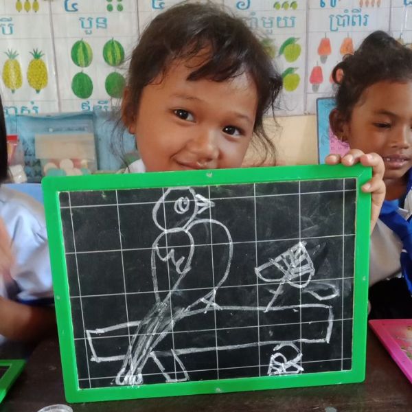A smiling preschool girl holds up a small green-framed chalkboard with a chalk drawing of a bird on a branch