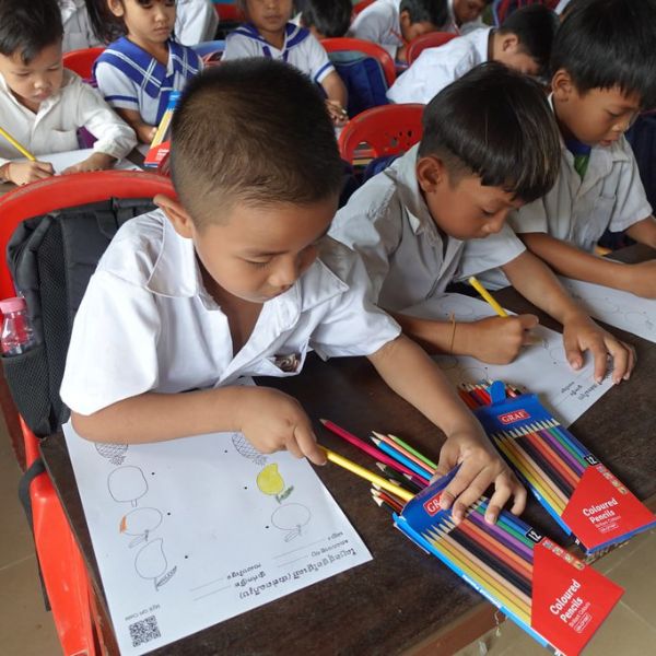 Two preschool boys in school uniform colour a worksheet with coloured pencils at a desk, with classmates behind them