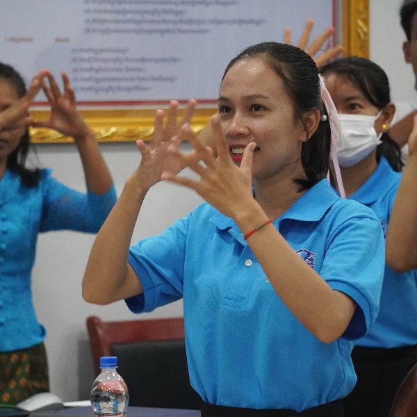 A teacher in a blue polo shirt makes a hand-shape gesture with both hands during a workshop activity