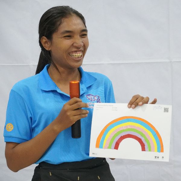 A smiling woman in a blue polo shirt holds a microphone and a sheet showing a colourful paper rainbow