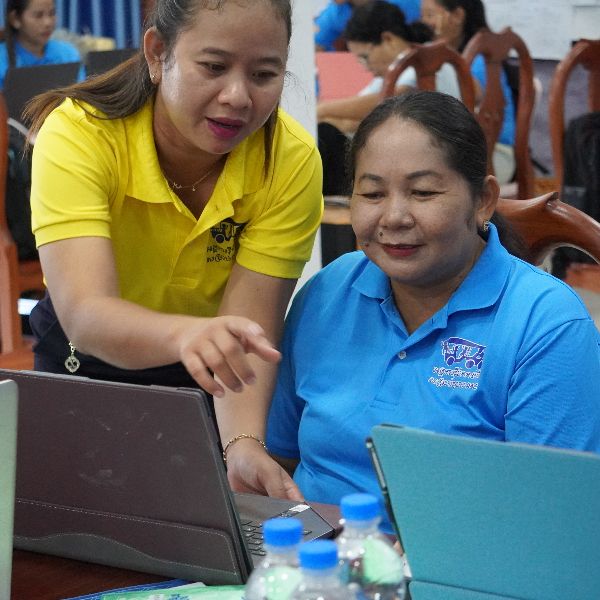 A woman in a yellow polo shirt leans over to point at a laptop screen beside a seated colleague