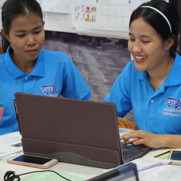 Two women in blue TukTuk polo shirts work together on a laptop at the workshop, one smiling