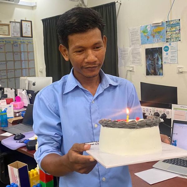 A man in a blue shirt carries a white birthday cake with a lit candle through the office