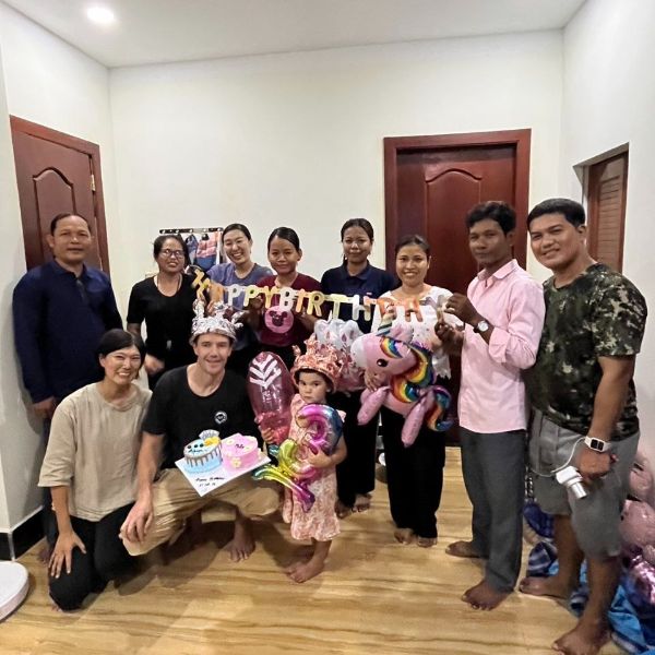 TukTuk staff, a visitor and a toddler in a party crown gather in a room with birthday balloons and cakes