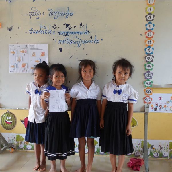 Four preschool girls in school uniforms stand in front of a classroom wall, one holding a worksheet