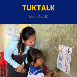TukTalk 80 cover: a preschool teacher helps a small girl in school uniform match picture cards on a classroom wall