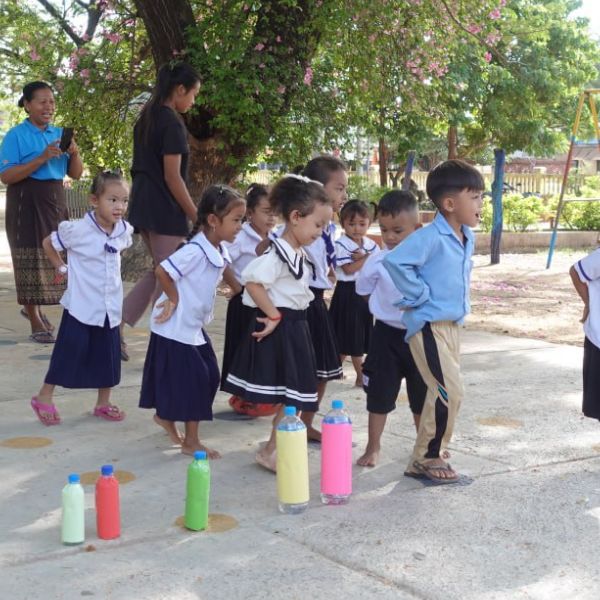 Preschool children in uniform stand in a playground in front of coloured bottles while teachers watch