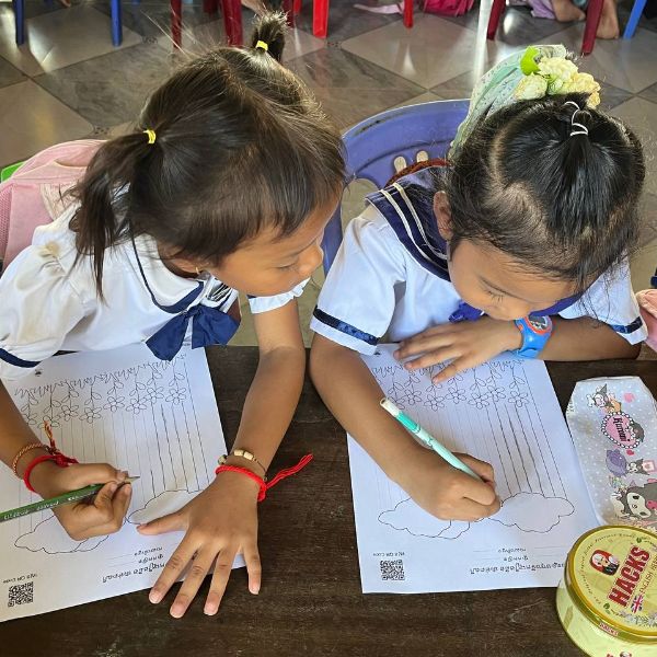 Two preschool girls draw with pens on worksheets of flowers and clouds at a wooden desk