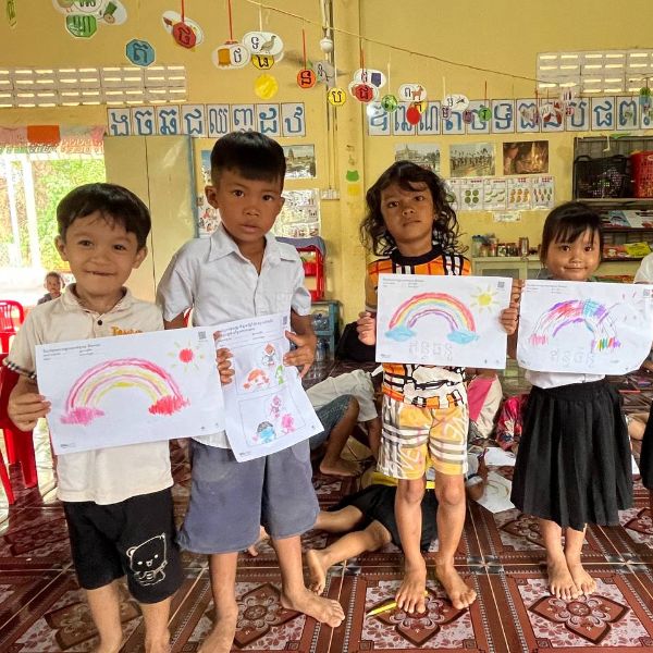 Four preschool children stand on a patterned mat in a classroom holding up their colourful rainbow drawings