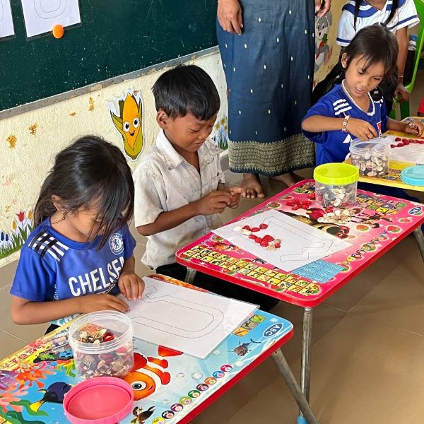 Preschool children sit at colourful low desks placing small red seeds onto outline worksheets