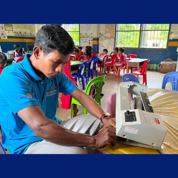 A man in a blue polo shirt operates a laminating machine in a preschool classroom with children behind