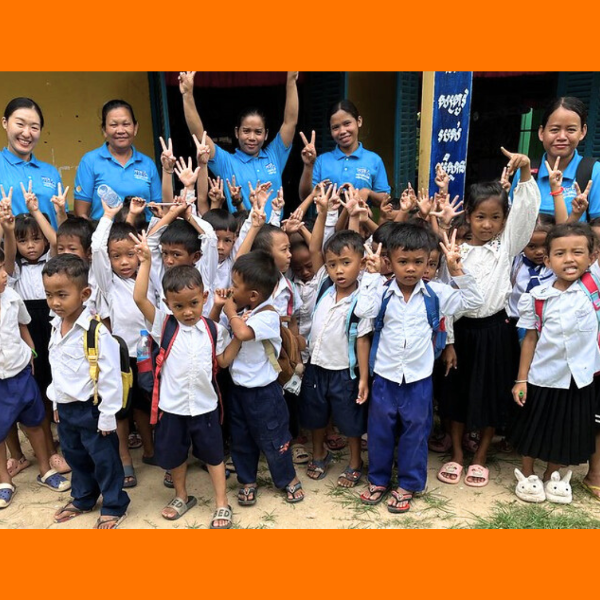 Preschool children in school uniforms raise their hands with peace signs, with teachers in blue polo shirts behind them
