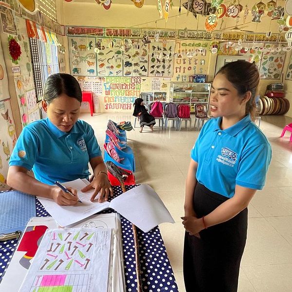 Two women in blue polo shirts in a preschool classroom, one seated writing on a worksheet, one standing