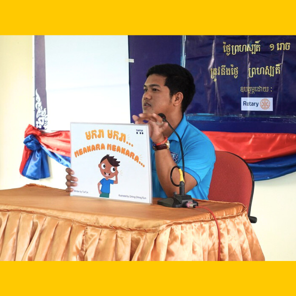 A presenter at a table with a microphone shows the picture book Meakara Meakara at the workshop