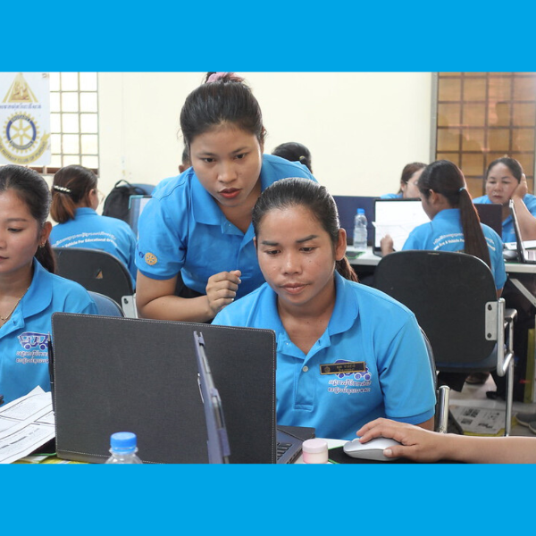 Teachers in blue polo shirts work at laptops during the computer lesson, one teacher helping another