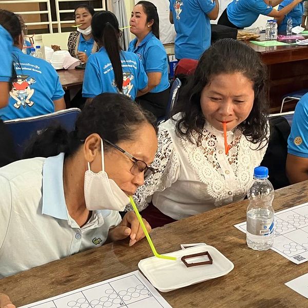 Two teachers at a workshop table use straws to move a small object, with others doing the same behind