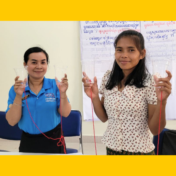 Two women each hold up plastic cups joined by red string, smiling in front of a flip chart