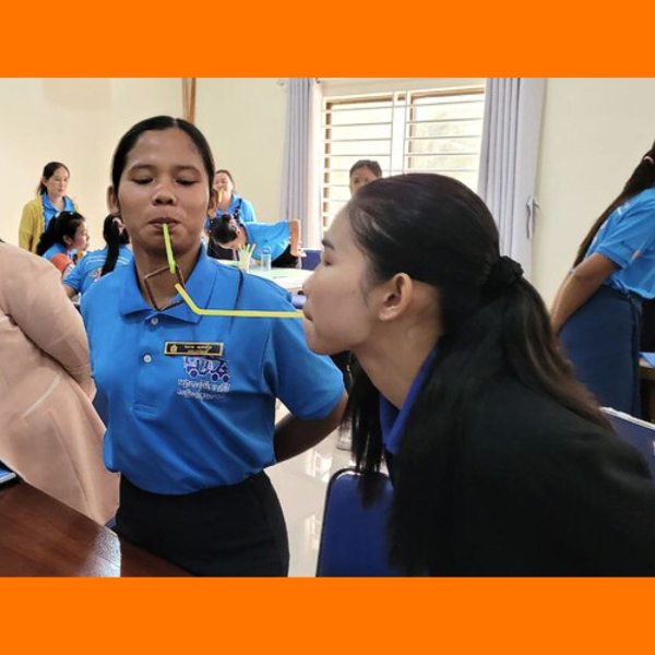 Two women in blue shirts face each other with a bendy straw between their mouths during a workshop game