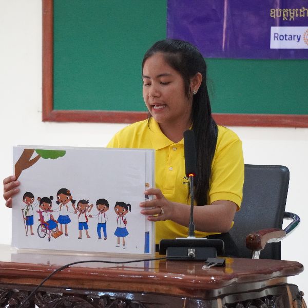 A woman in a yellow polo shirt sits at a microphone showing a picture of cartoon schoolchildren