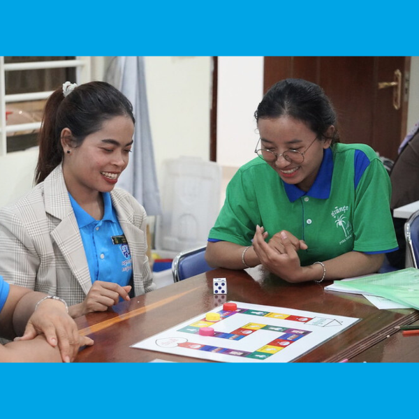 Two women in polo shirts laugh as they play a colourful board game with a dice at a table