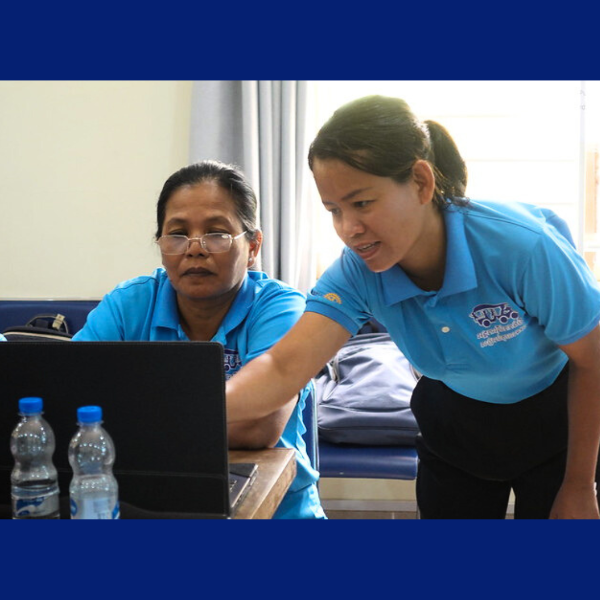 A woman in a blue polo shirt leans over to help a seated colleague with a laptop at the workshop