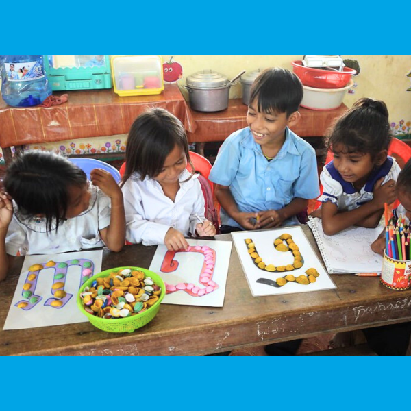 Children decorate Khmer letter worksheets with bottle caps at a classroom table