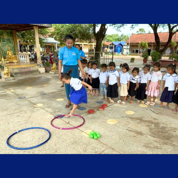 A child hops between hoops in an outdoor game while a TukTuk staff member and a line of preschoolers watch