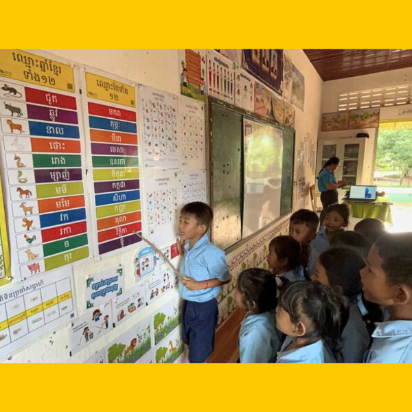 A child points at a Khmer chart on the classroom wall while classmates watch
