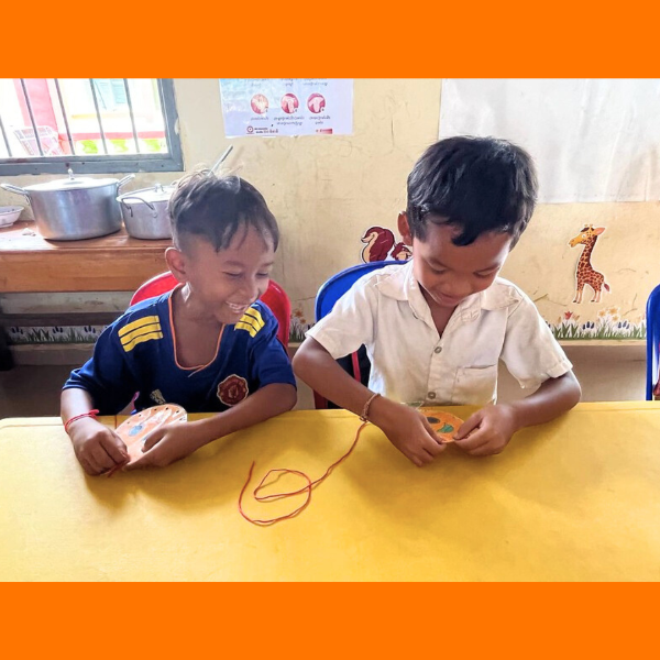 Two boys smile as they play with string and paper craft cards at a classroom table