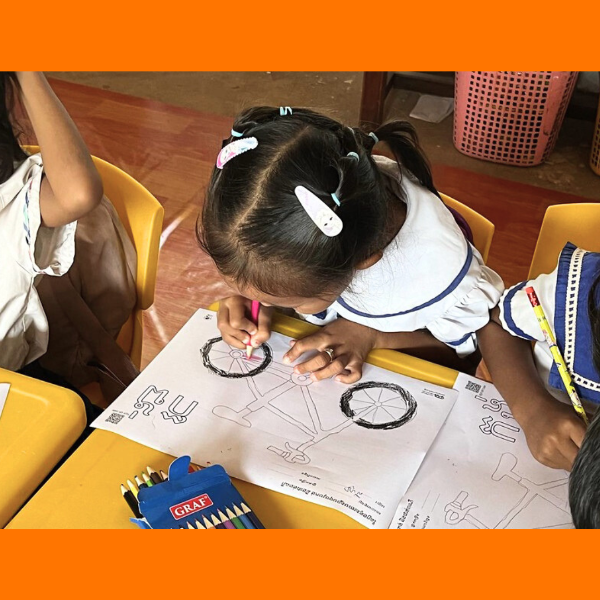 A girl colours a bicycle worksheet with a pink pencil at a classroom table