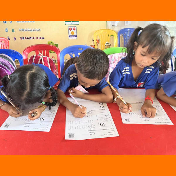 Three children write Khmer letters on practice worksheets at a red classroom table