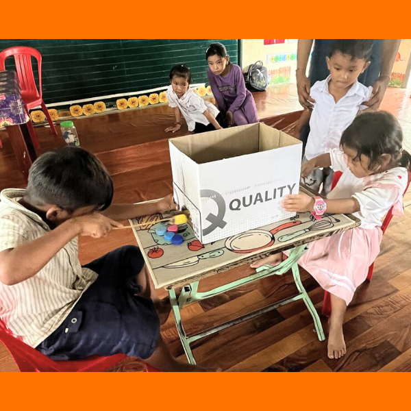 Children play a table game with a cardboard box and colourful pieces on the classroom floor