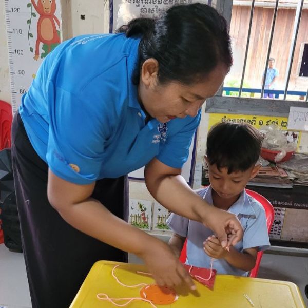 A TukTuk staff member helps a child thread string through a paper craft at a classroom table