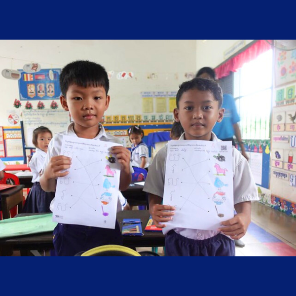 Two boys proudly hold up their matching worksheets, coloured in with pencils, in class