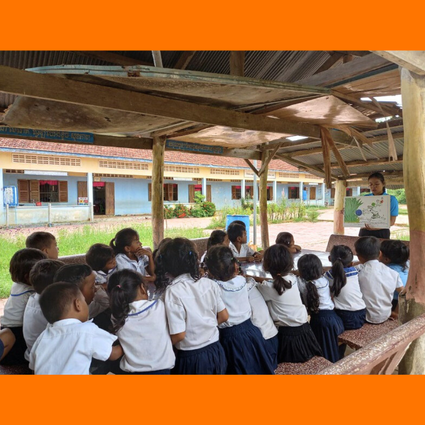 A teacher shows a picture book to preschool children sitting in an open-air shelter at school