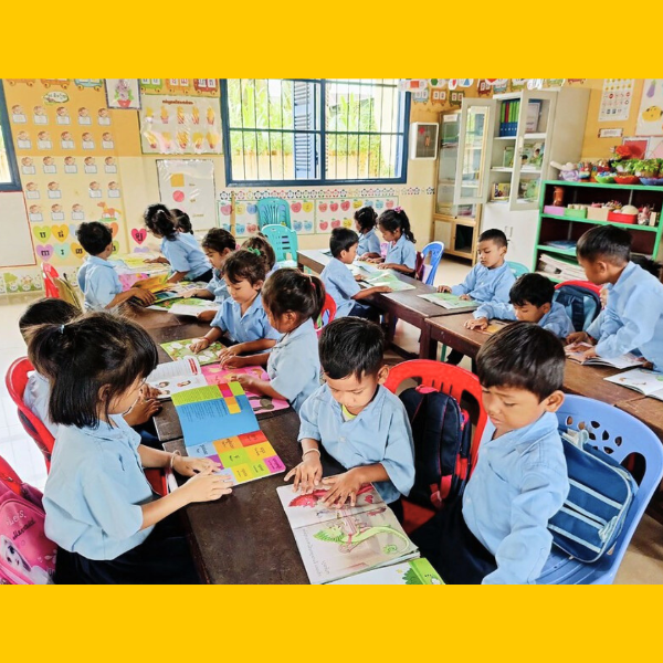 Children in light blue uniforms read picture books at long classroom tables