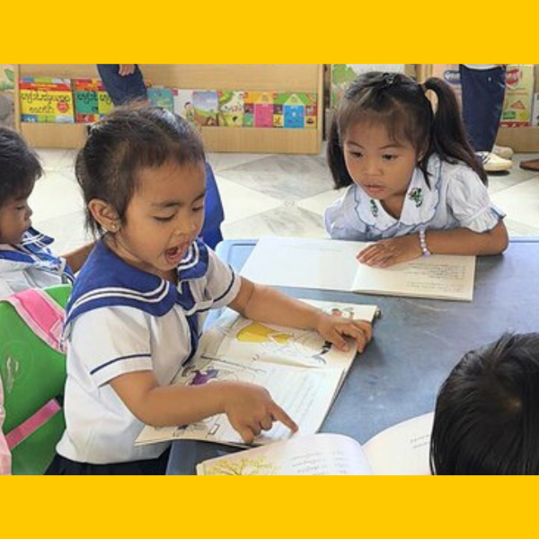 Two girls read a picture book at a table, one pointing at the page