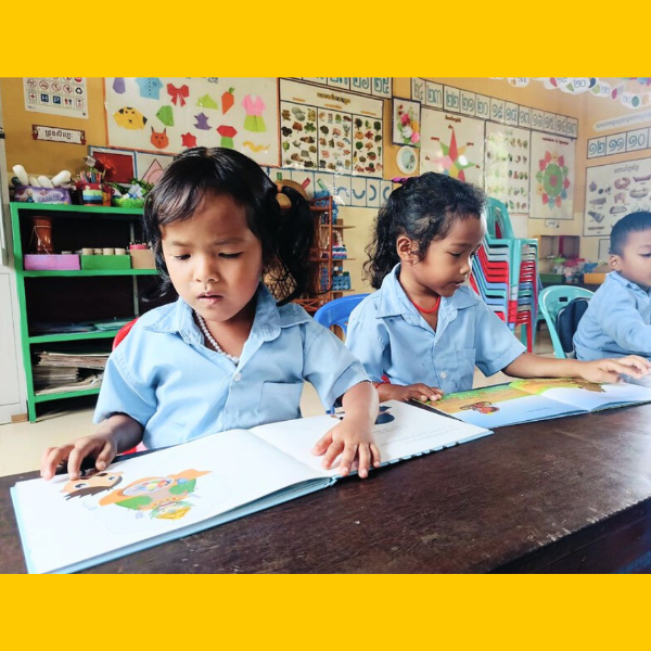 A girl turns the pages of a large picture book at a classroom desk