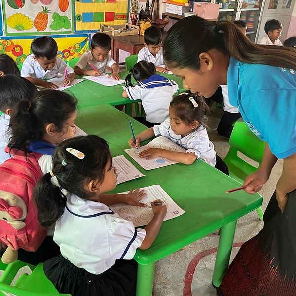 A TukTuk staff member leans over children colouring worksheets at green classroom tables
