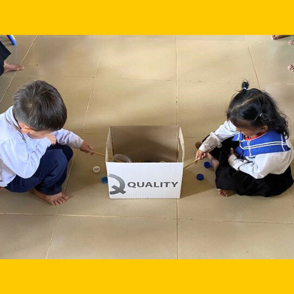 Two children use sticks to move bottle caps around a cardboard box on the classroom floor