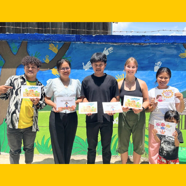 Volunteers and TukTuk staff hold up picture-book pages in front of the colourful mural wall